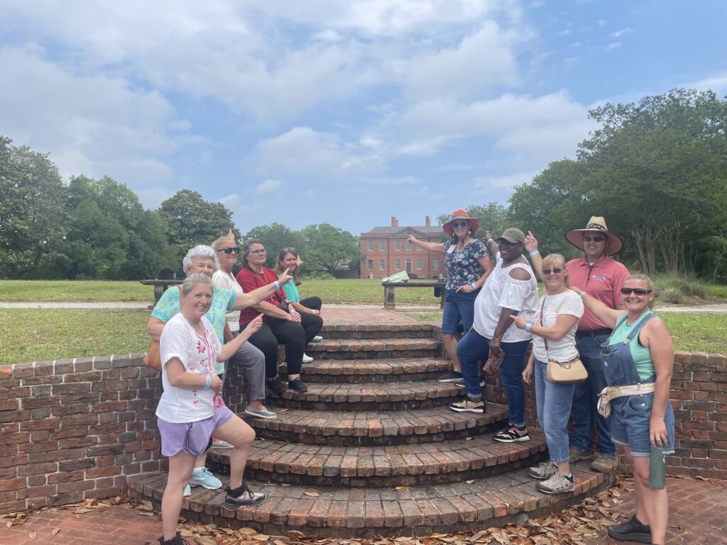 The 2025 Extension Master Gardener class graduates smiling and pointing toward Tryon Palace during a field trip in New Bern, NC.